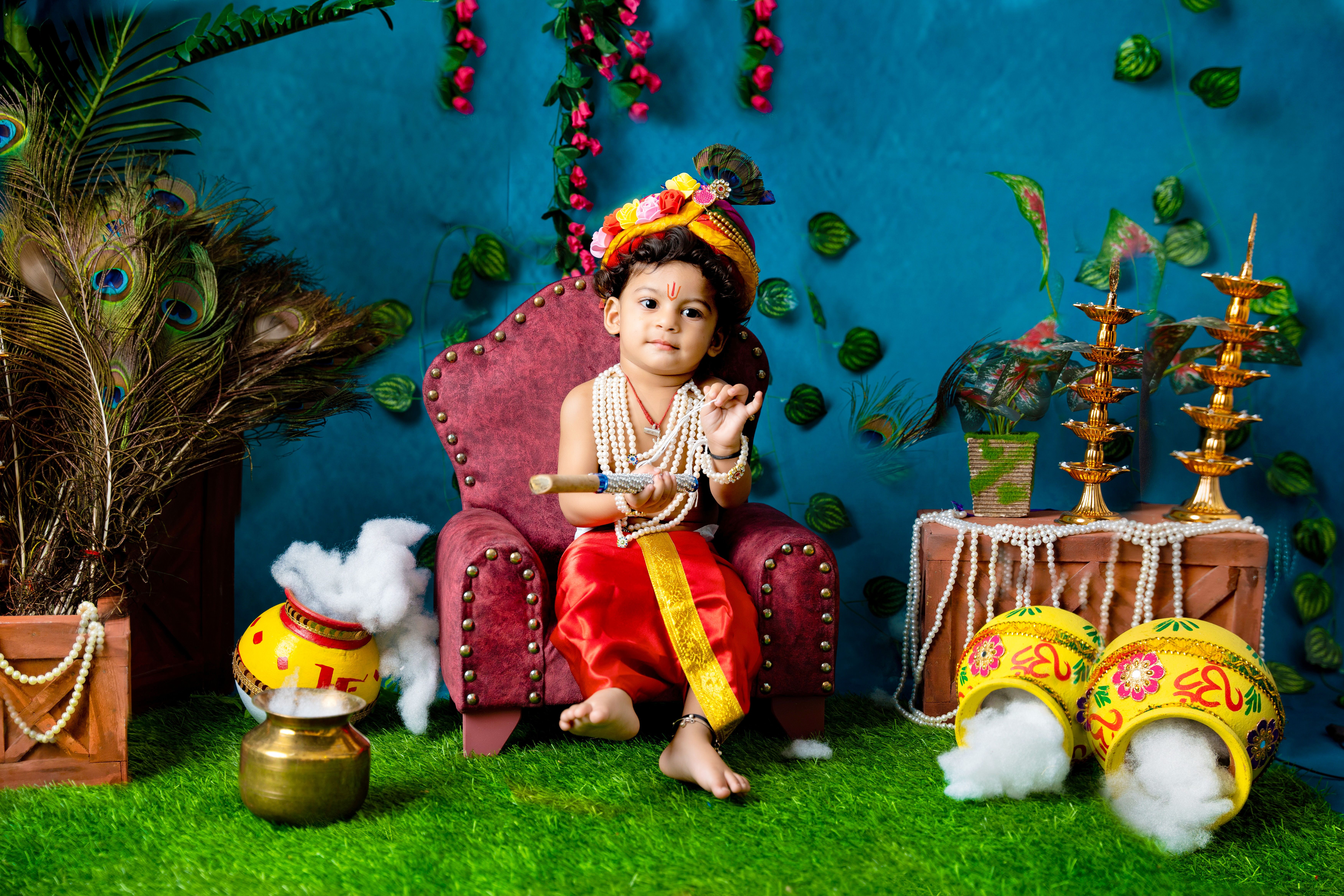 A young child dressed as Lord Krishna, holding a flute, in a colorful Indian traditional setting.