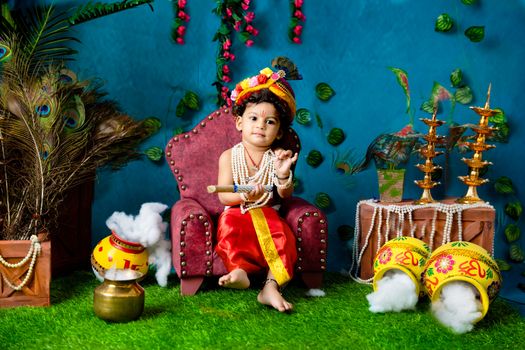 A young child dressed as Lord Krishna, holding a flute, in a colorful Indian traditional setting.