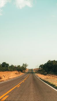A lone asphalt road stretches through rural countryside with dry grass under a summer sky.