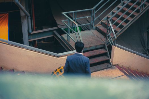 A man in a suit is seen from behind walking up a metal staircase in an urban setting.