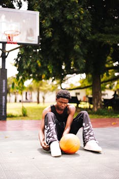 A young man in sportswear ties his shoe on a sunny outdoor basketball court.