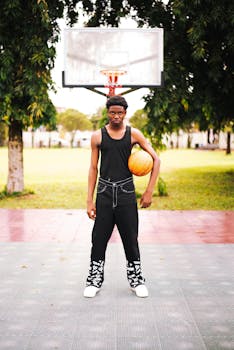 A young man stands with a basketball on an outdoor court surrounded by trees.