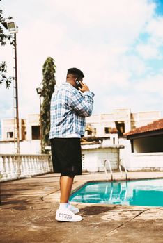 A man stands by an outdoor pool, talking on his smartphone on a sunny day.