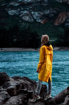 A woman stands on rocky terrain overlooking a serene river, dressed in a bright yellow raincoat.
