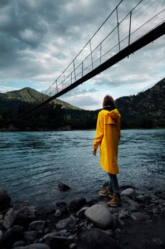 Woman in yellow raincoat standing by the river under a suspension bridge.