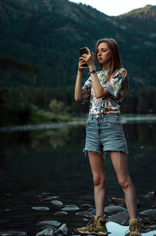 A Woman Standing by the Lake in Mountains and Taking a Picture with Her Phone 