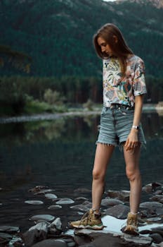A young woman stands by a tranquil mountain lake, enjoying a summer day outdoors.