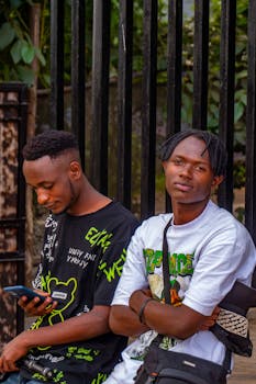 Two young men standing by a fence in casual outfits, one looking at a smartphone, outdoors.