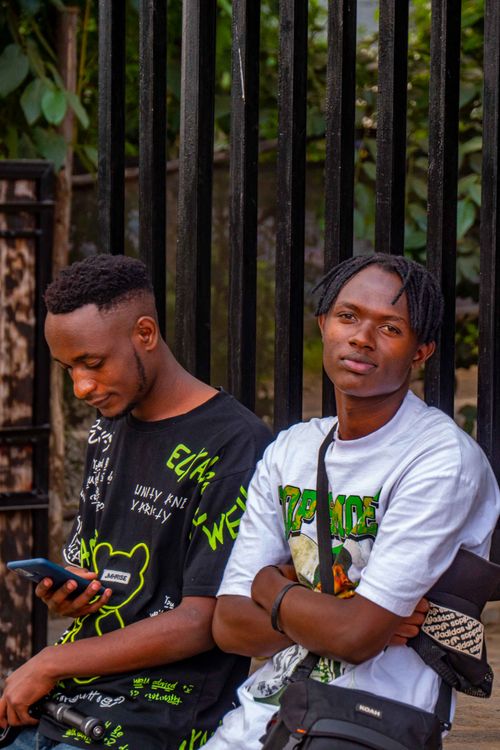 Free Two young men standing by a fence in casual outfits, one looking at a smartphone, outdoors. Stock Photo