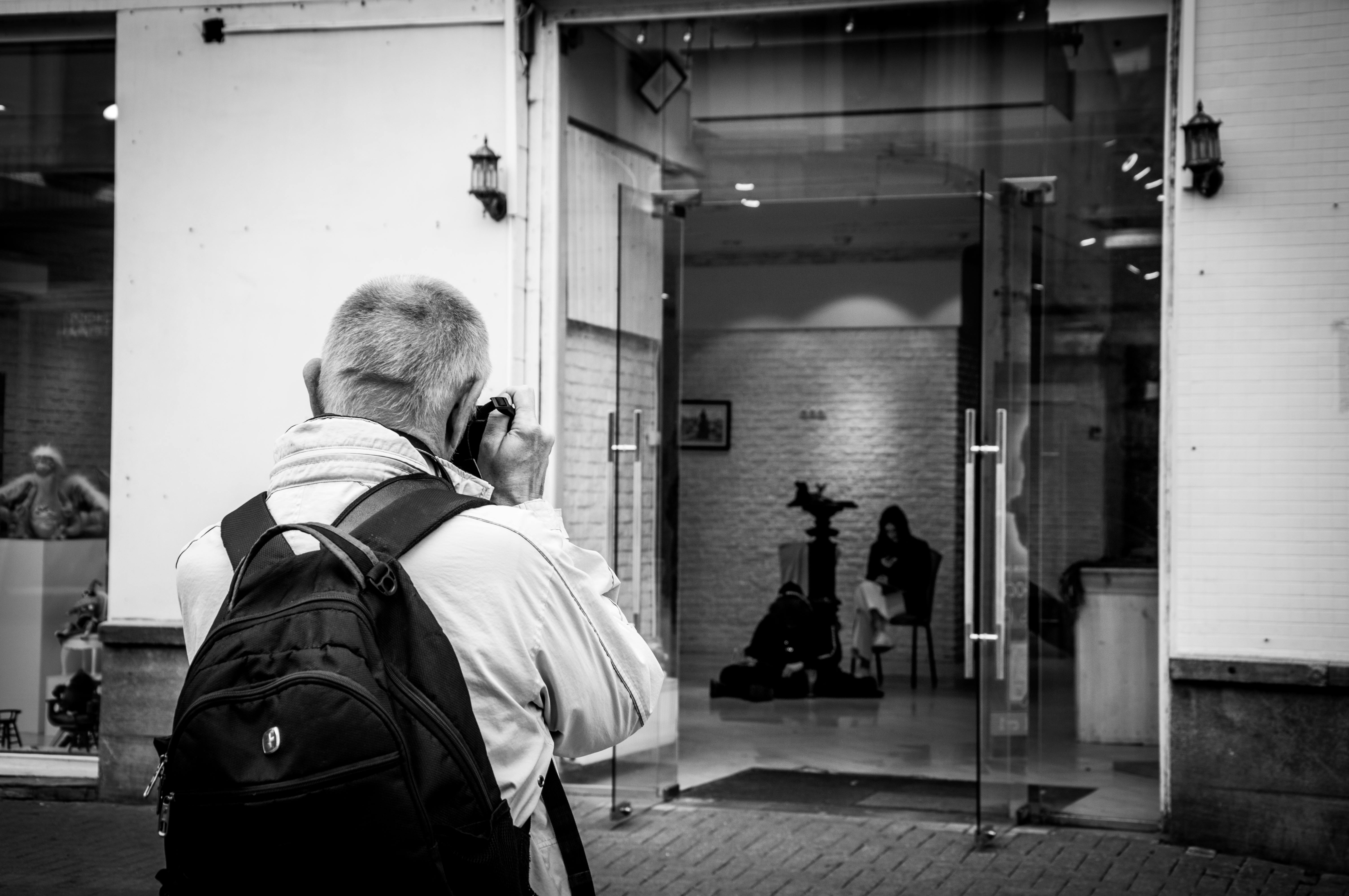 Black and white image of a photographer with a backpack taking a picture in an urban setting.