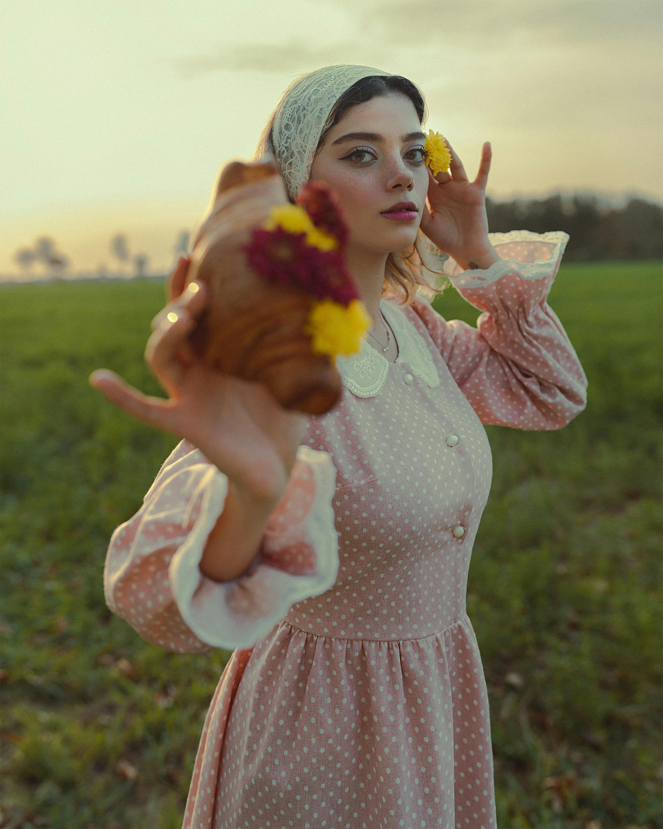 Whimsical photo of a woman in a vintage pastel dress holding a croissant adorned with flowers at sunset.