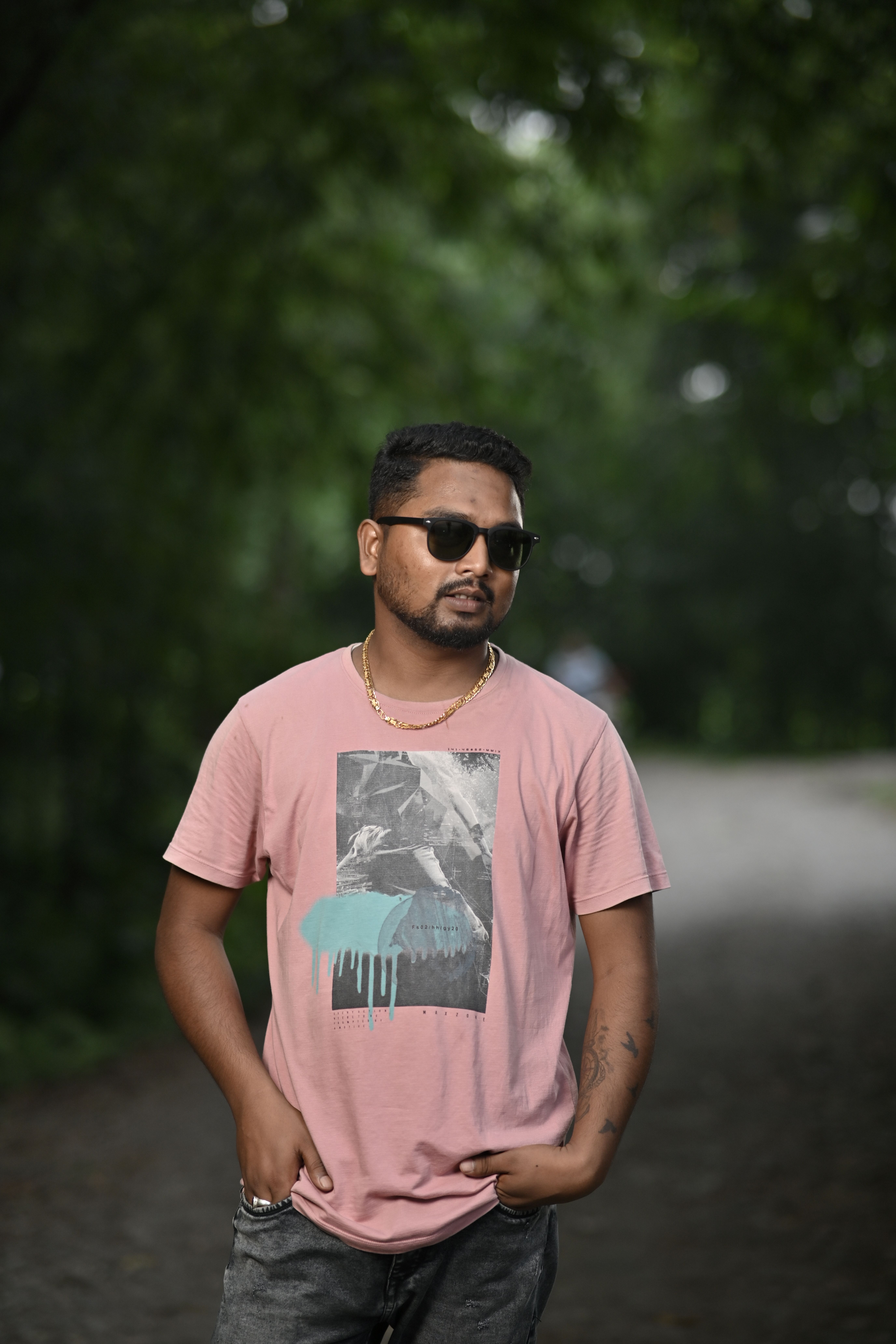 Man in pink graphic t-shirt and sunglasses standing on a wooded path.