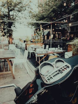 Outdoor restaurant patio with tables and a motor scooter foregrounded in an urban setting.