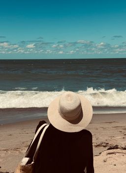 Back view of a woman in a straw hat sitting on a sandy beach facing ocean waves.