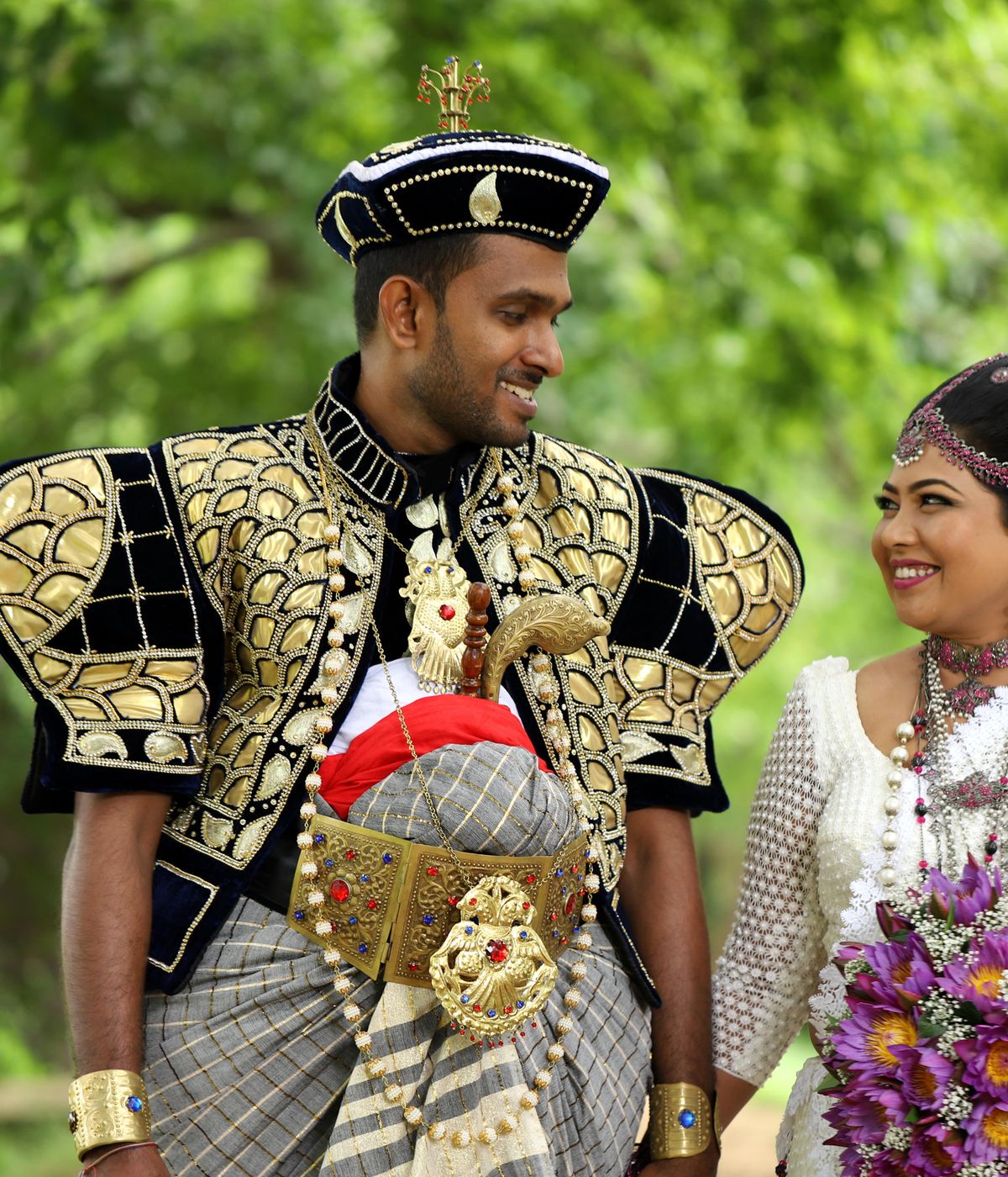 Laughing Sri Lankan couple — groom in black and gold Kandyan nilame regalia, bride with orchid bouquet