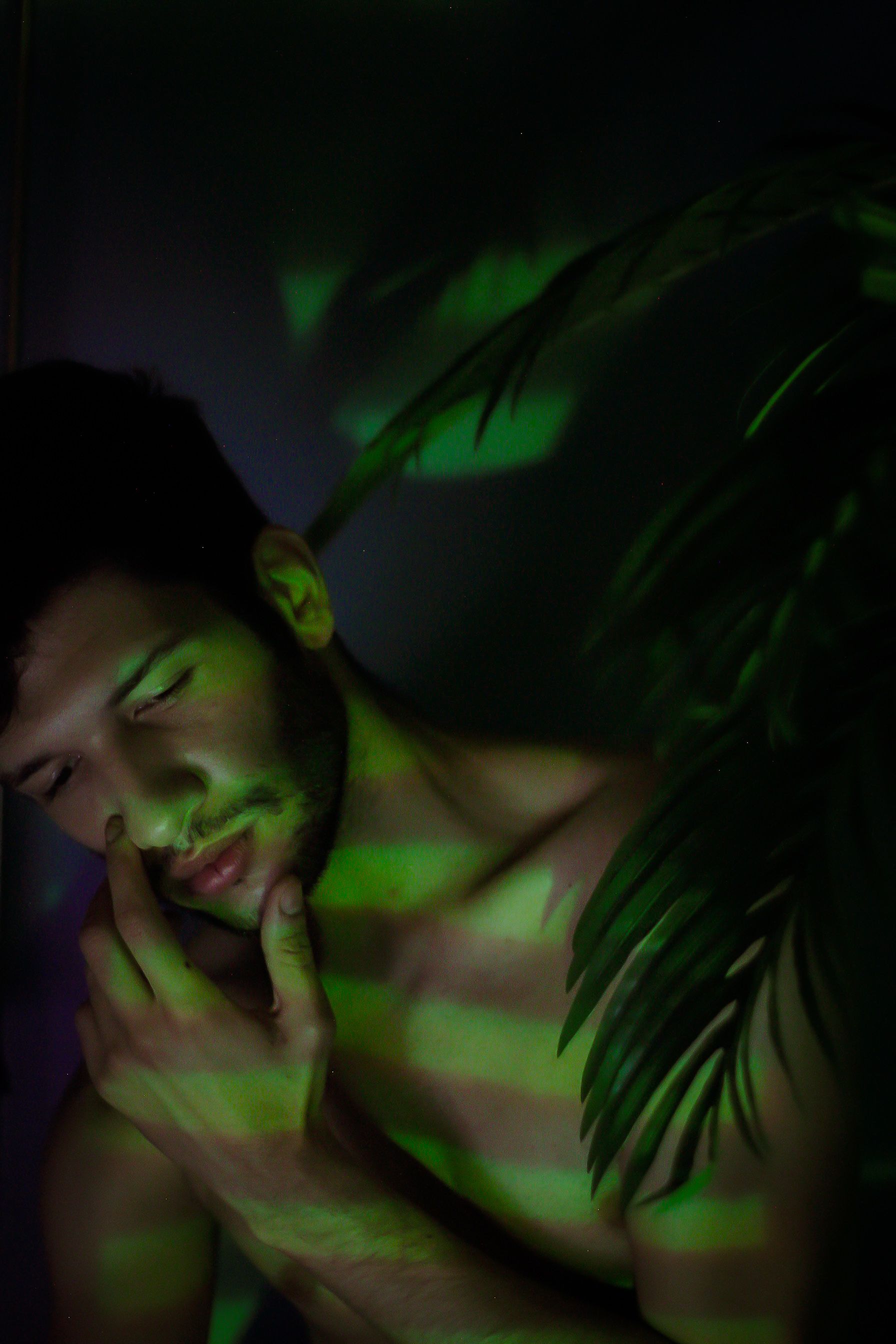 A contemplative shirtless man, eyes closed, enveloped in moody green shadows with plants.