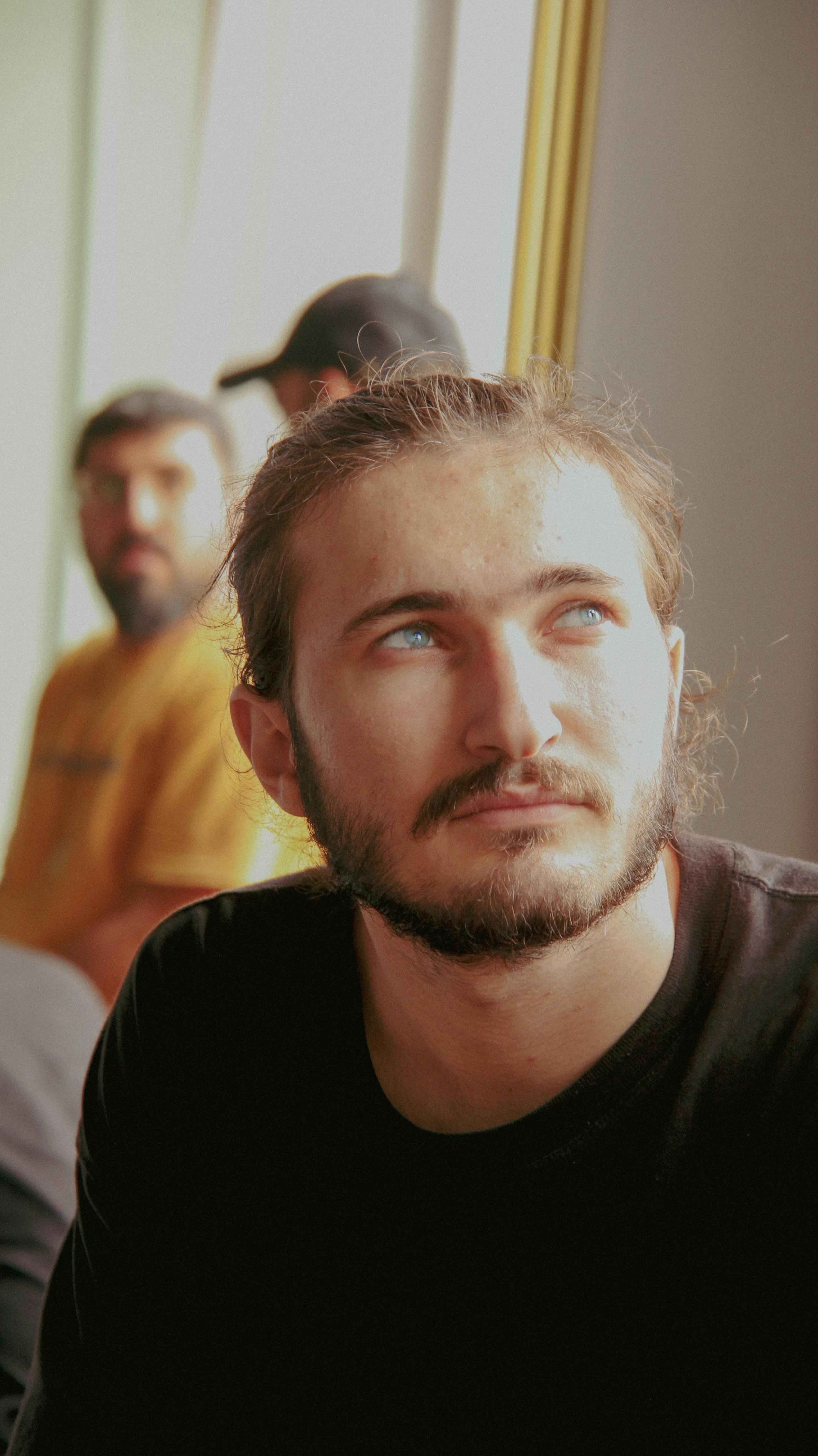 A thoughtful young man with a beard looking upwards in a sunlit indoor room.
