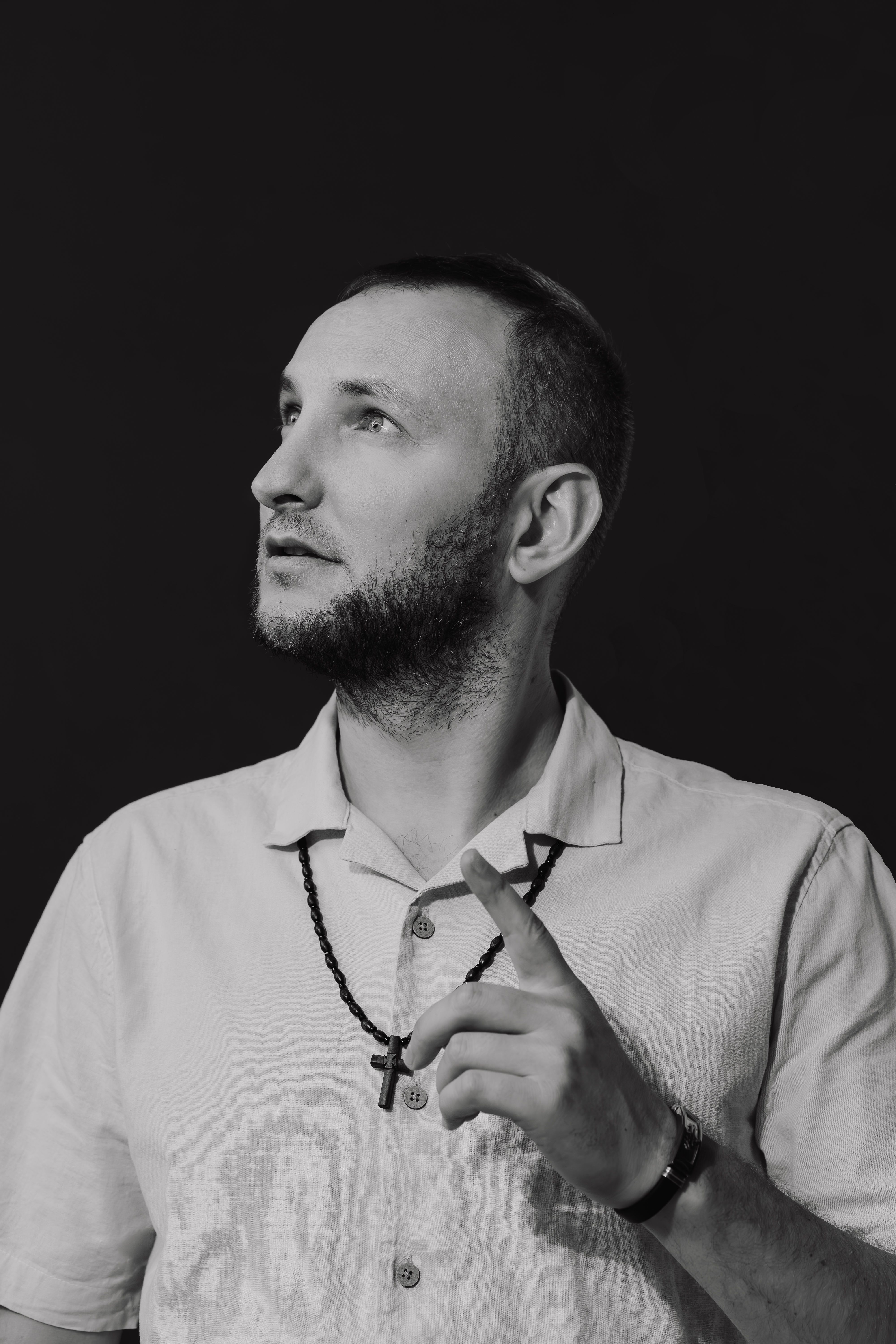 A pensive man in a white shirt and necklace, gazing upward in black and white portrait.