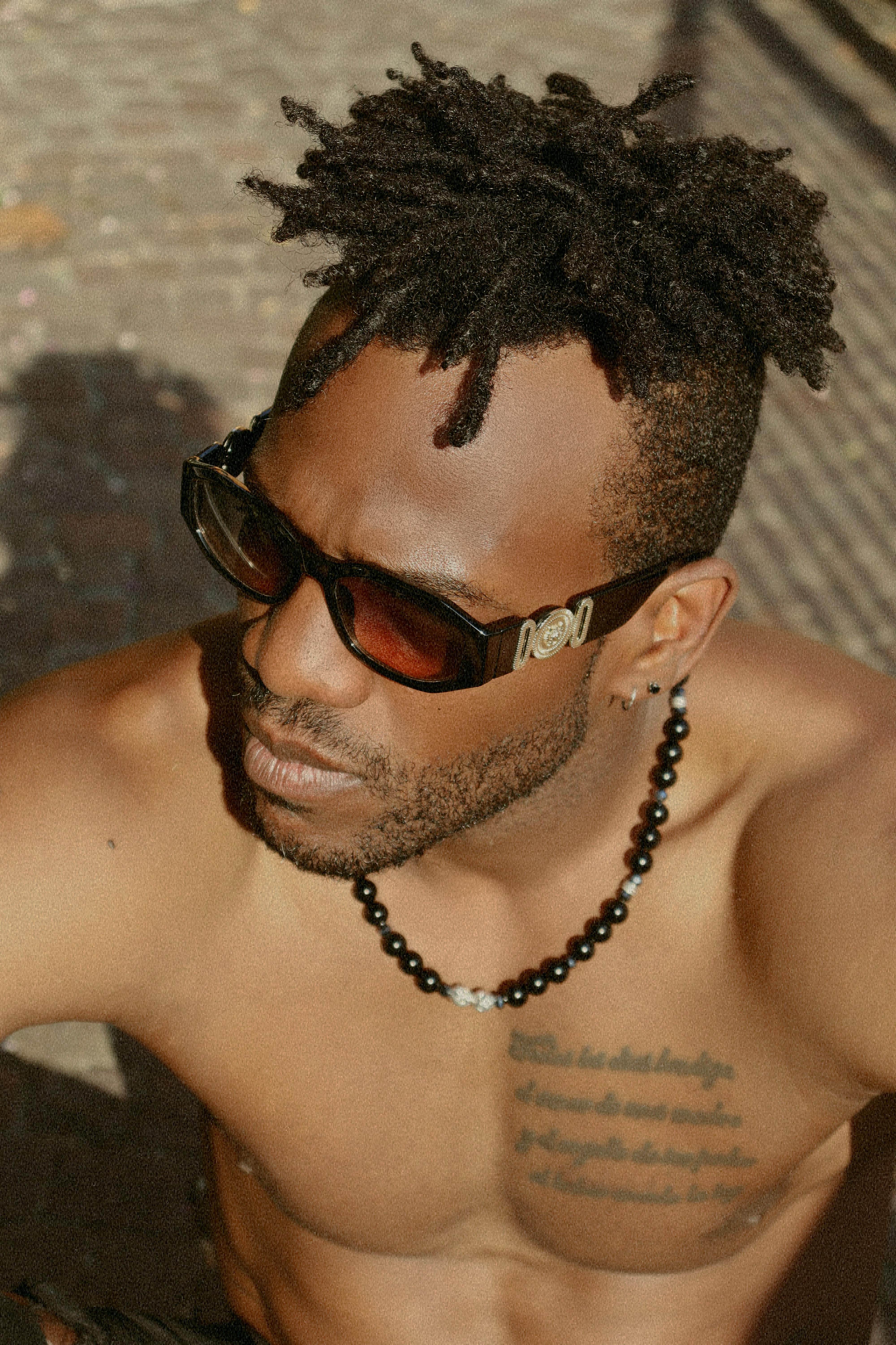 A shirtless man with dreadlocks, sunglasses, and tattoos in a bright outdoor setting.
