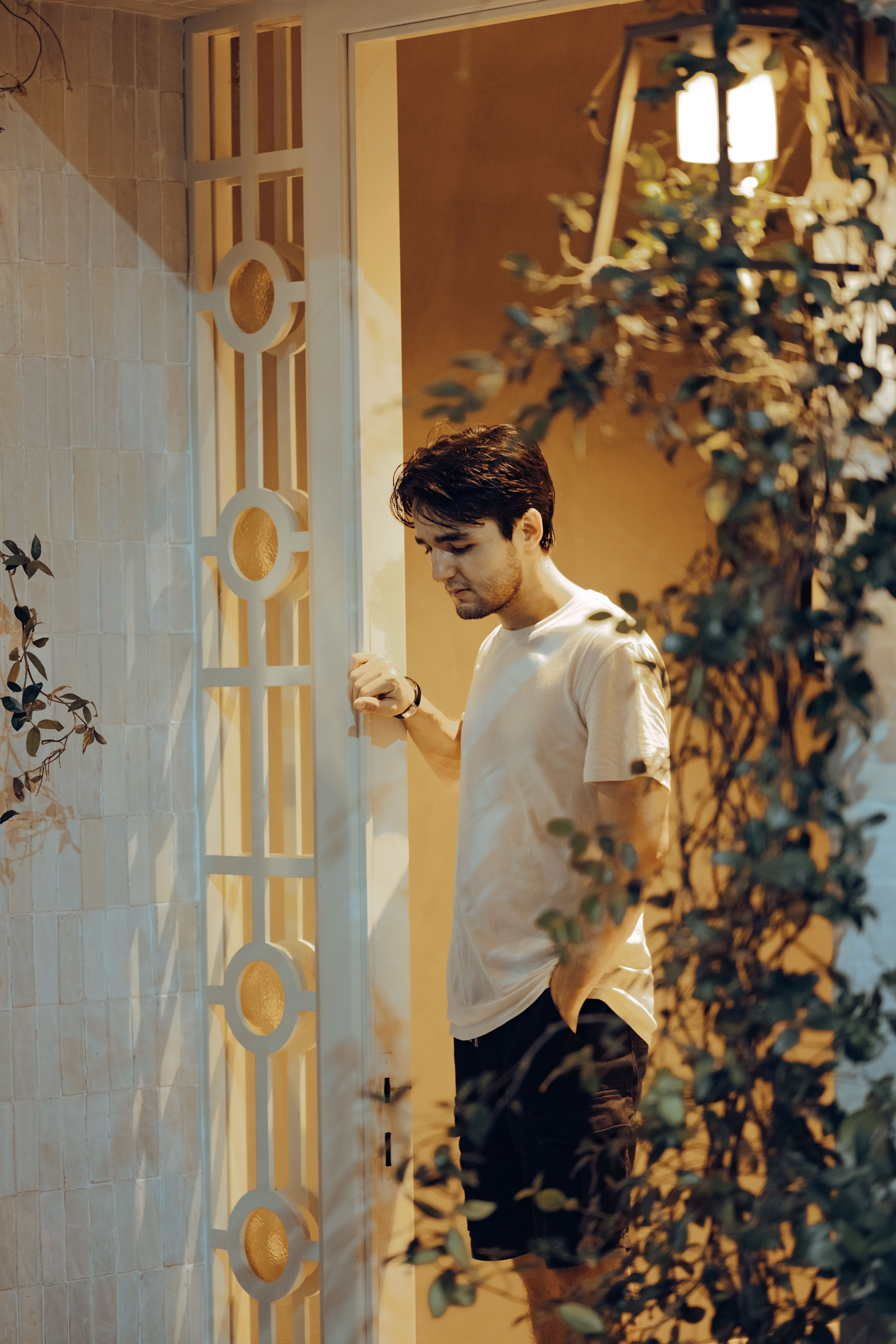 A thoughtful man stands in a doorway at night, surrounded by soft light and greenery.