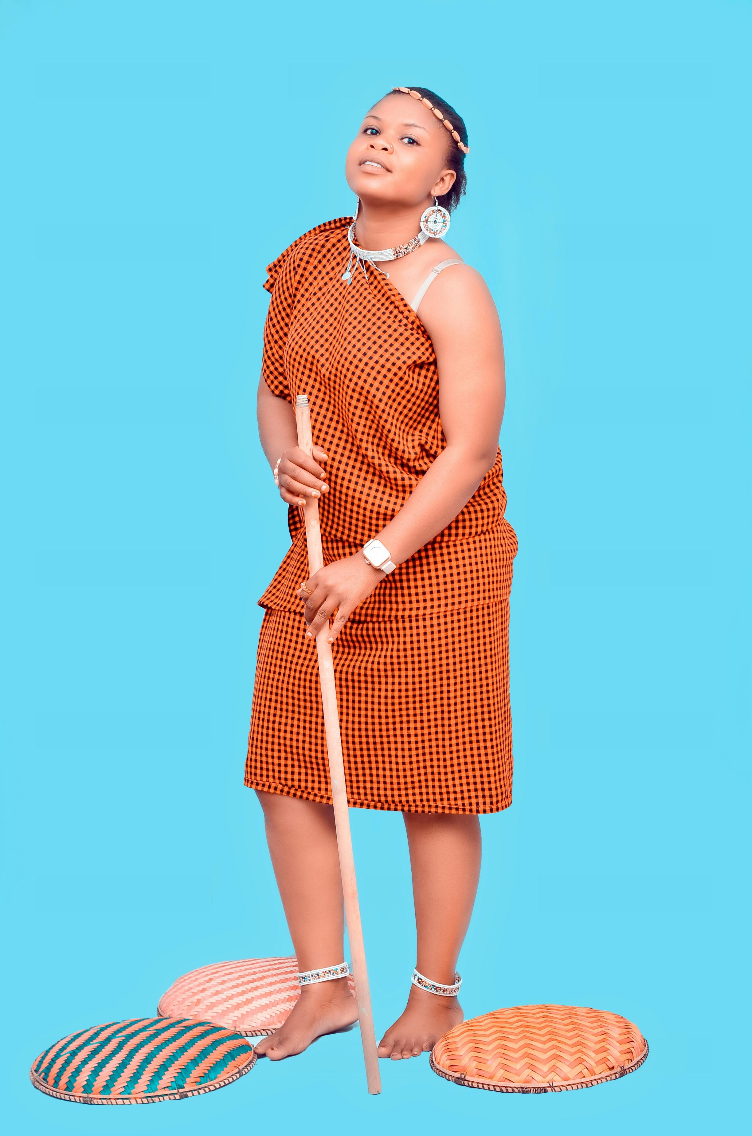 African woman in traditional attire posing with a wooden stick in a studio setting.