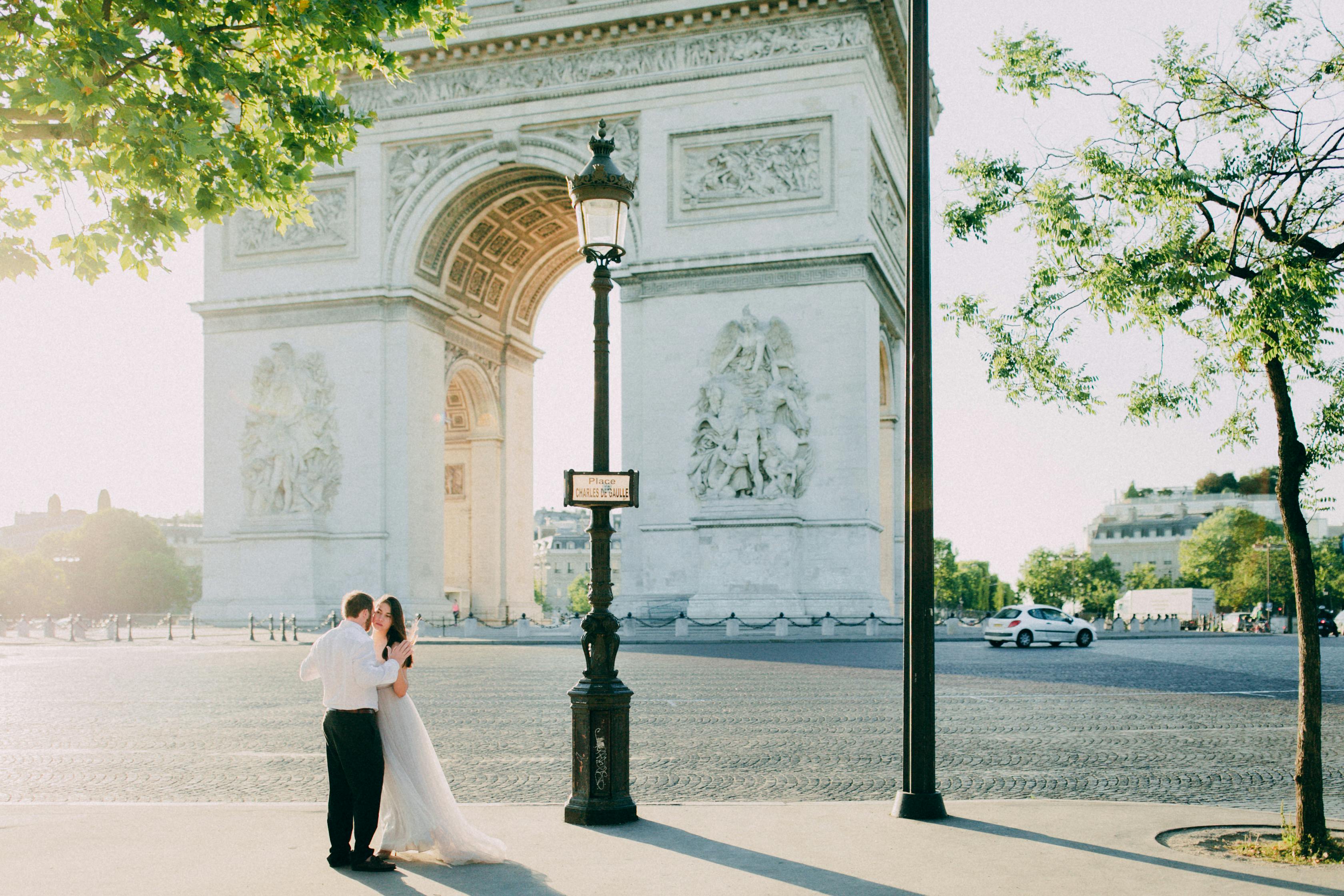A couple in wedding attire embraces by the Arc de Triomphe in Paris on a sunny day.