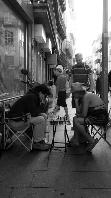 Black and white shot of people playing chess on a busy urban sidewalk, capturing city life. — Xadrez, Portugal.