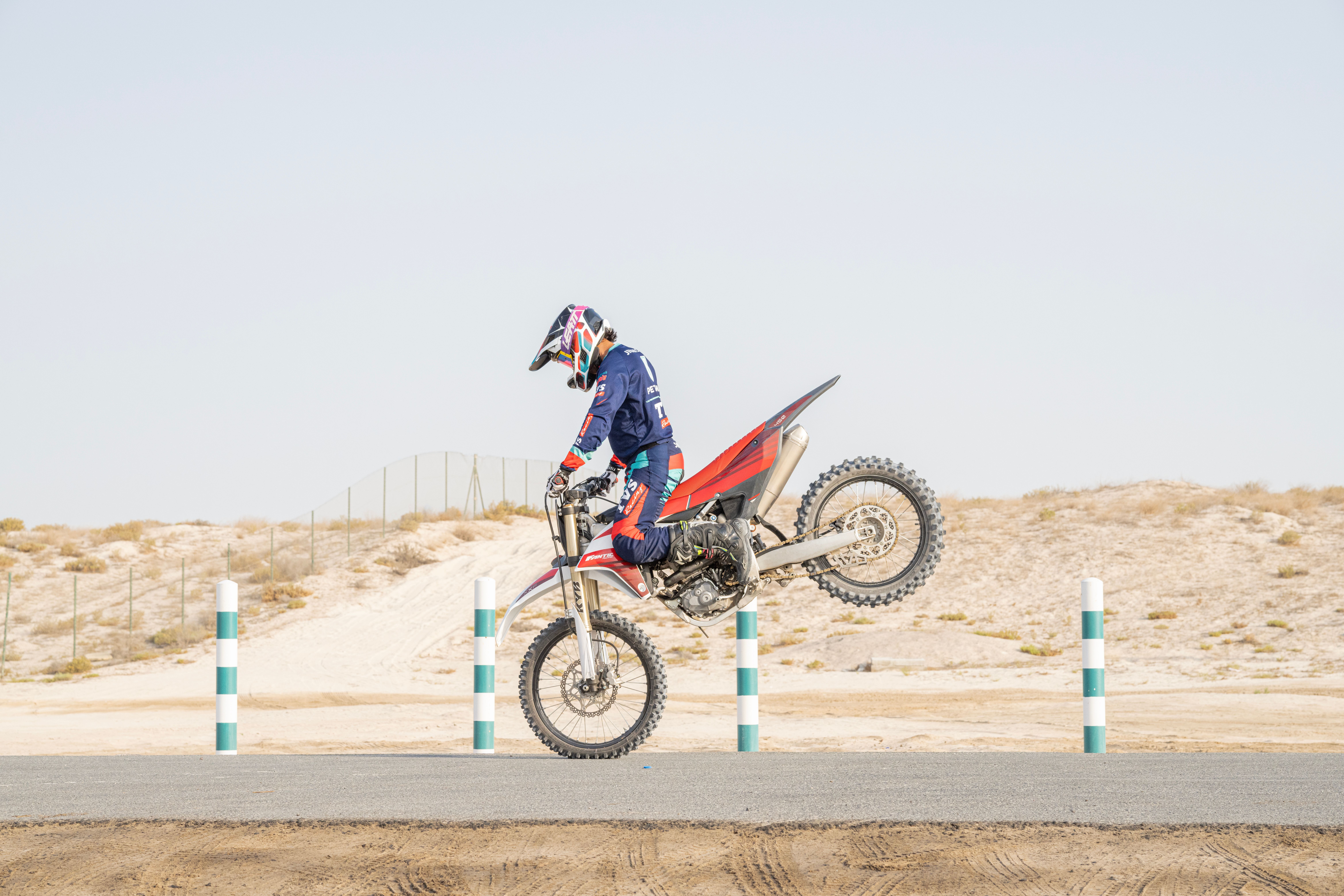 A motorcyclist performs a high jump in a desert landscape, kicking up dust.