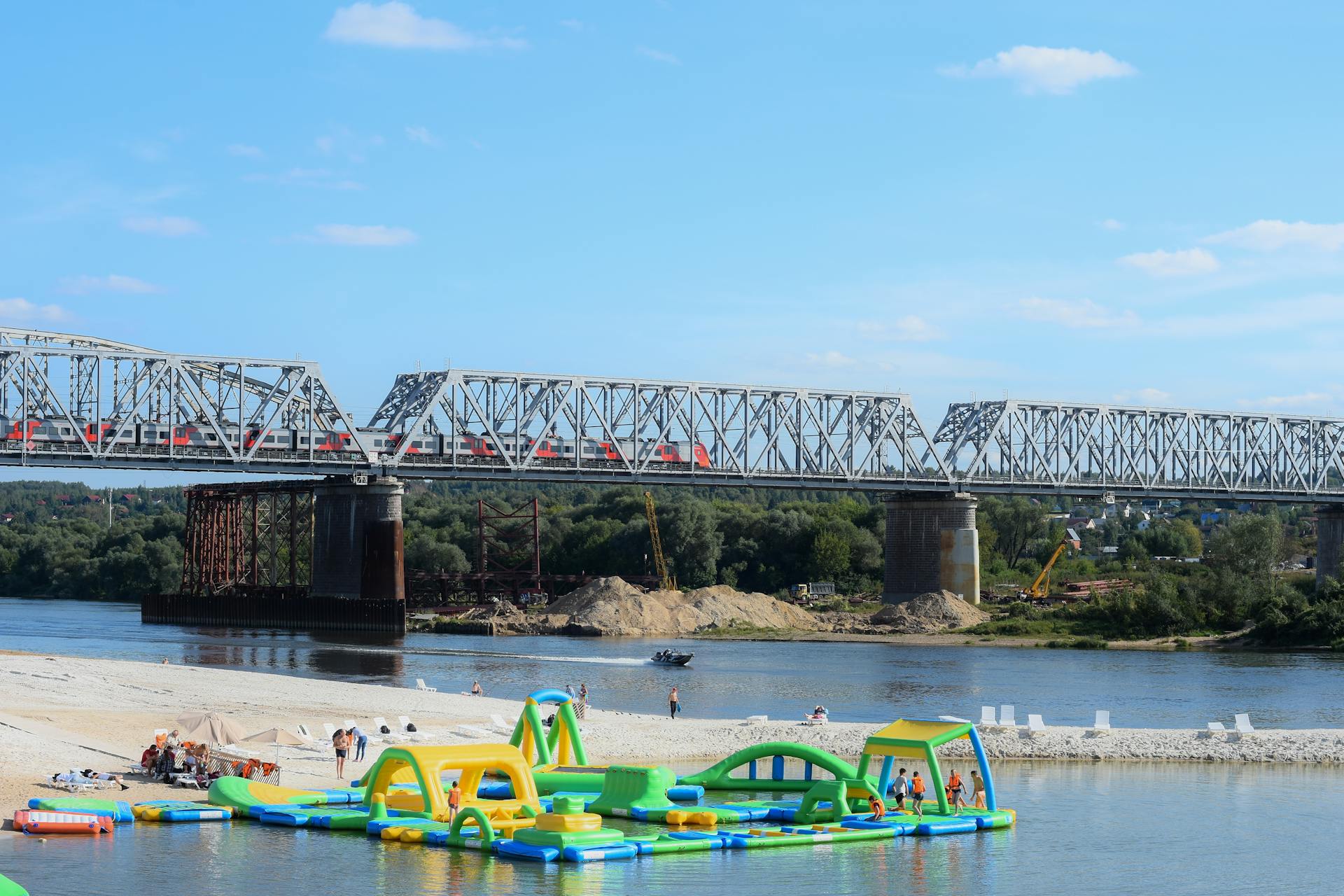 Scenic river view with a truss bridge, beach, and floating water park on a sunny day.