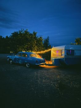 Atmospheric evening shot of a blue vintage car and lit camper in a rural setting.