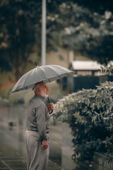 Senior man holding an umbrella standing on a wet sidewalk during rainfall.
