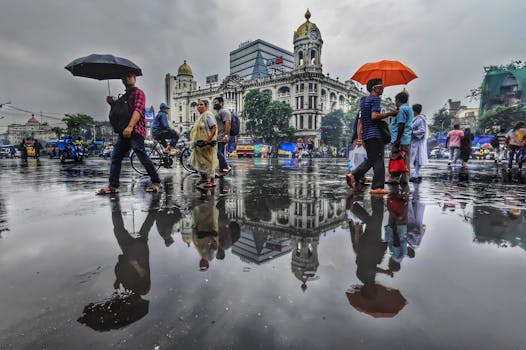People walking with umbrellas near the famous Metropolitan Building in Kolkata, India with reflections in rain puddles.