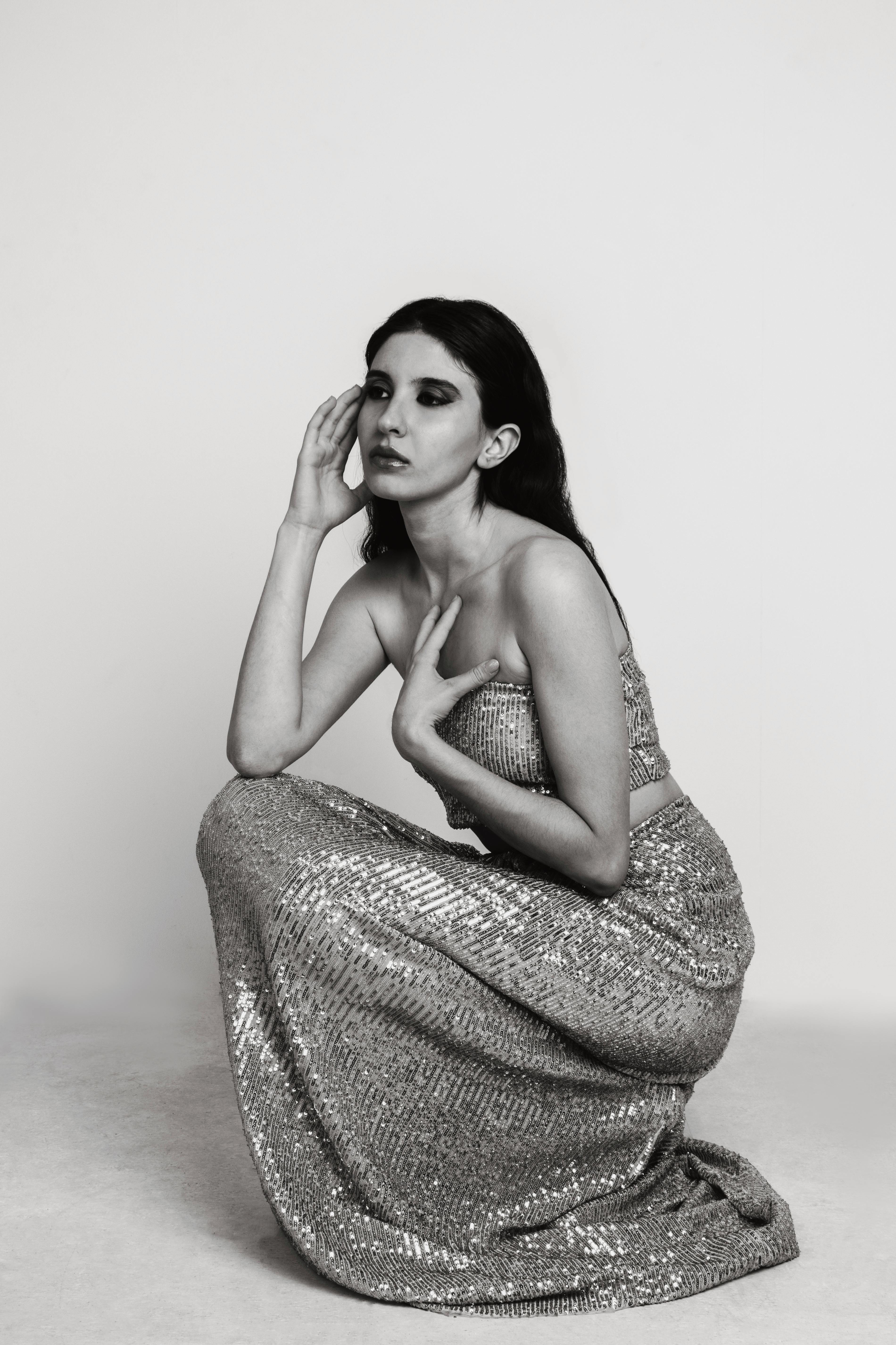 A glamorous woman kneels gracefully, showcasing a sparkling sequin gown, in a monochrome studio portrait.