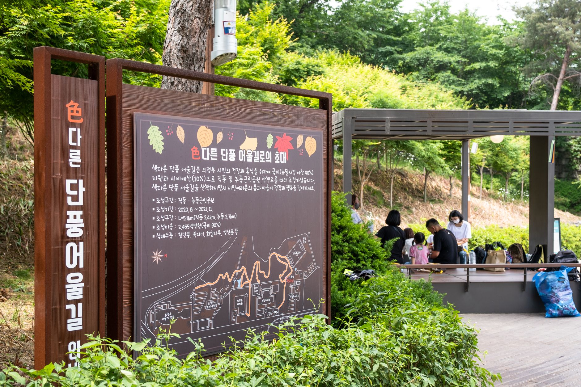 Outdoor park scene with a detailed map signboard and visitors relaxing under a pavilion.