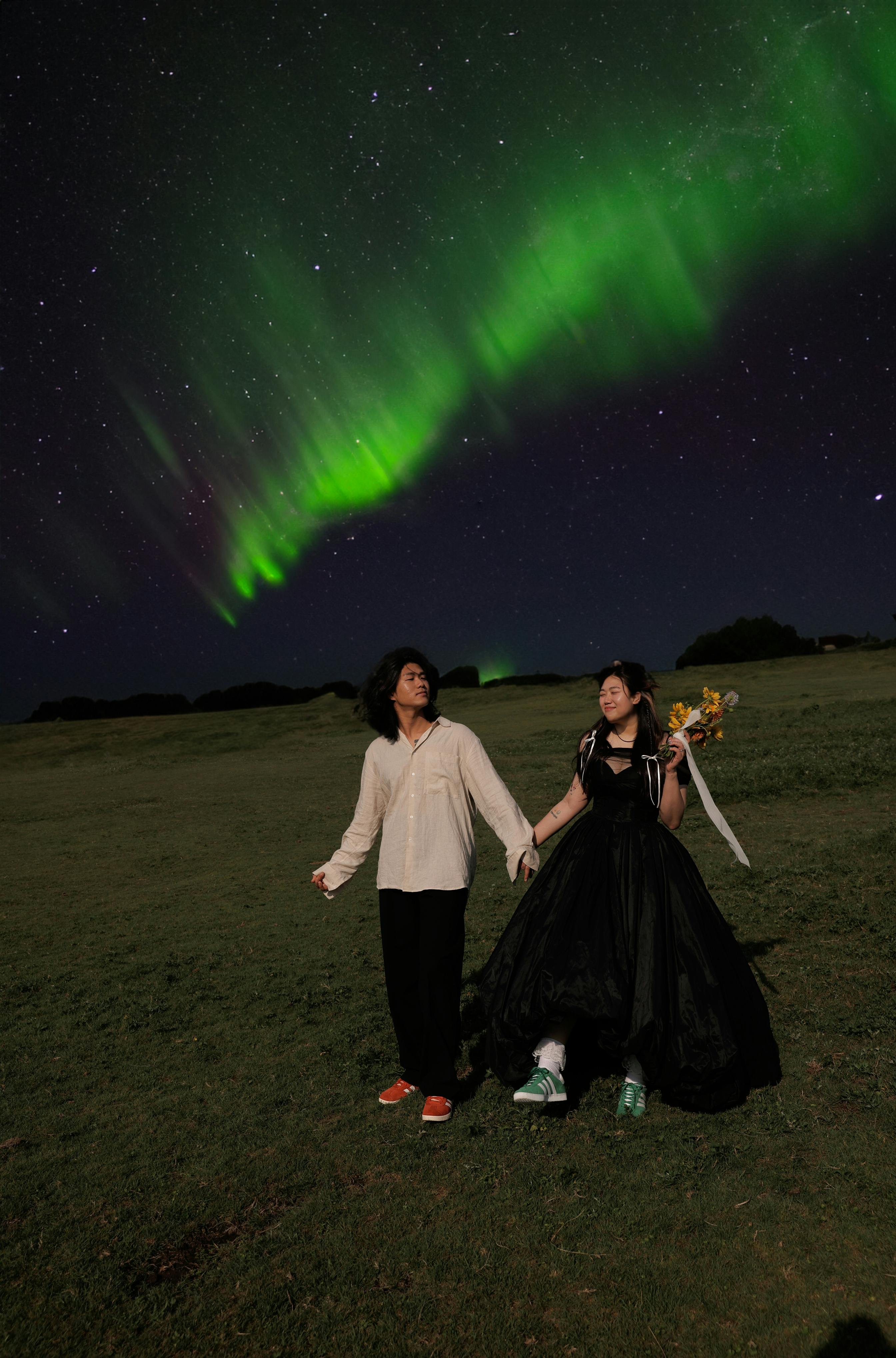 A couple holds hands beneath the vibrant northern lights in a grassy field at night.