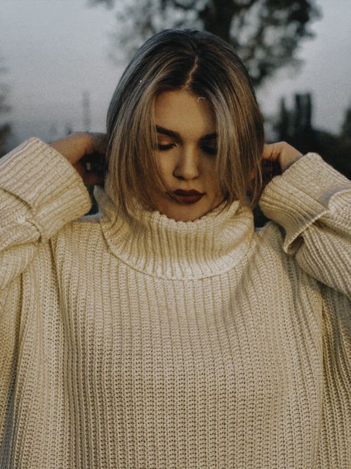Free Blonde woman adjusting white turtleneck sweater outdoors with soft, dramatic lighting. Stock Photo
