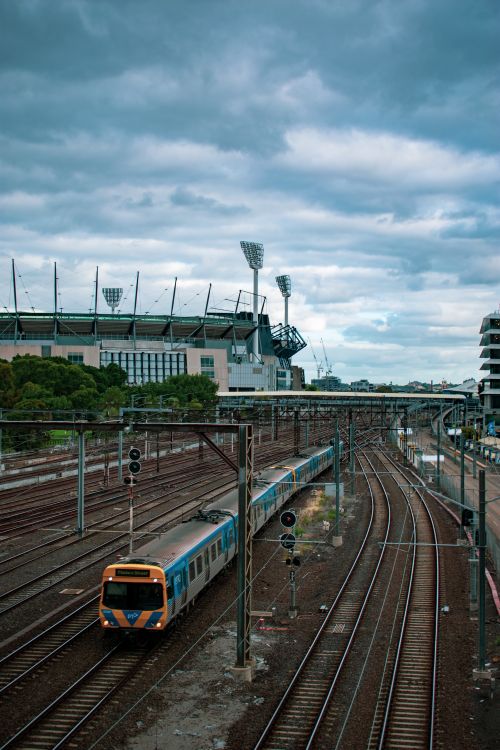 Free Overhead view of a passenger train on railway tracks near a large stadium under cloudy skies. Stock Photo