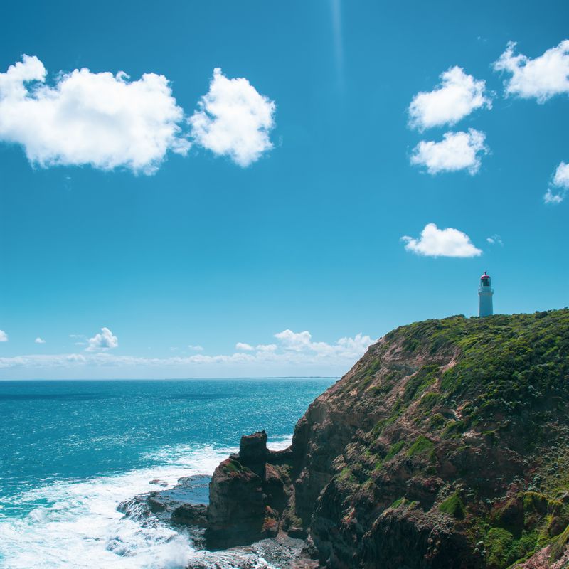 A small white lighthouse on a green cliff above a rocky shore and blue sea