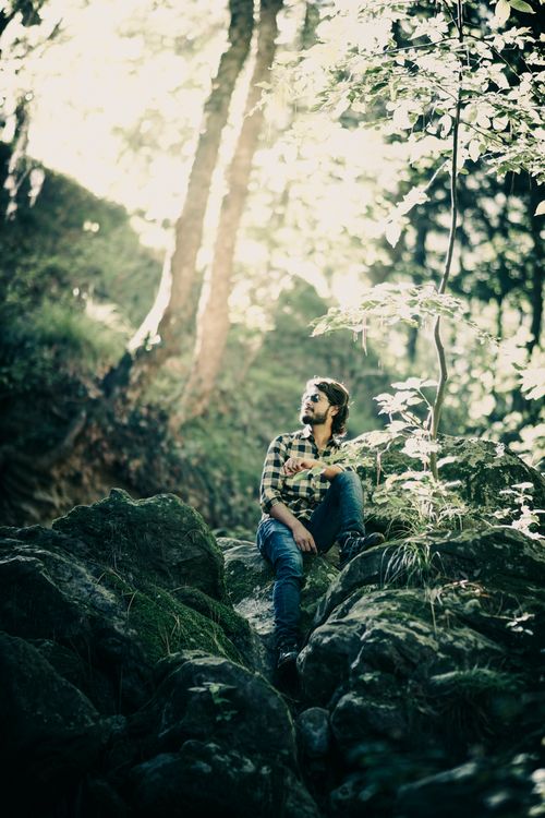 Free Caucasian man relaxing on rocks in forest sunlight, wearing sunglasses and checked shirt. Stock Photo