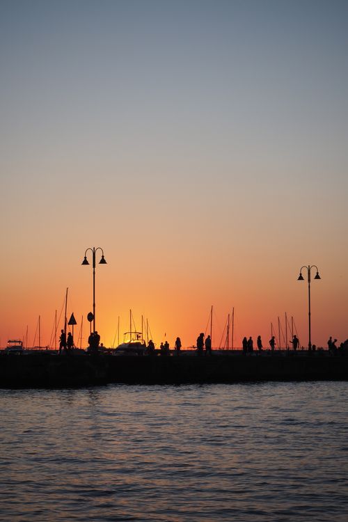 Free Captivating sunset over a bustling pier with silhouetted figures and lamp posts against the sea. Stock Photo