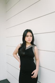 Stylish portrait of a young woman in a black dress against a white wall, showcasing elegance and contemporary fashion.