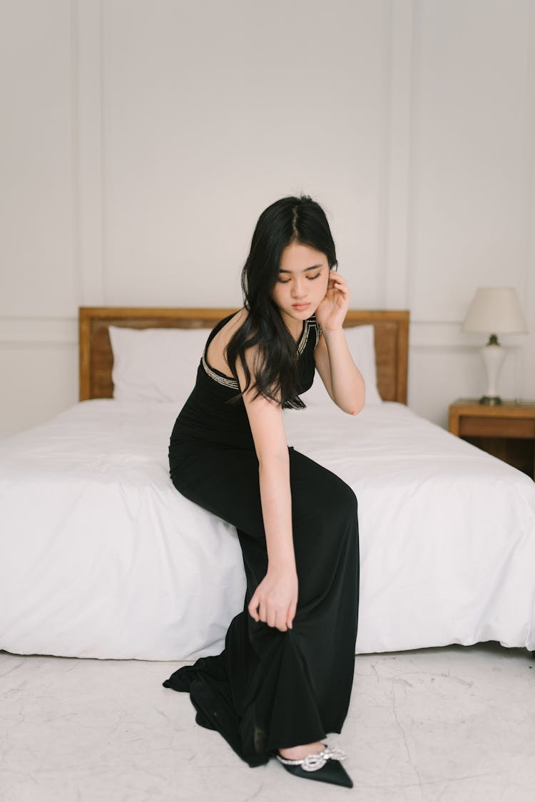 Young Woman In A Black Dress Sitting On The Bed 