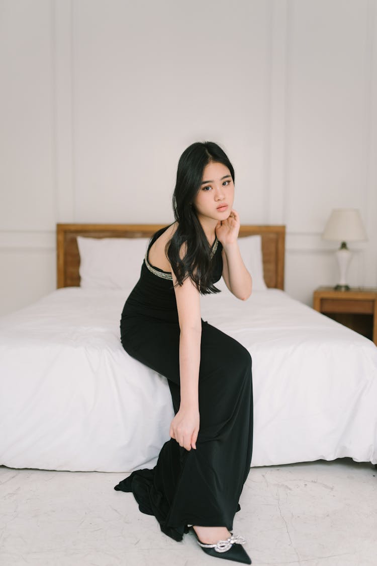 Young Woman In A Black Dress Sitting On The Bed 