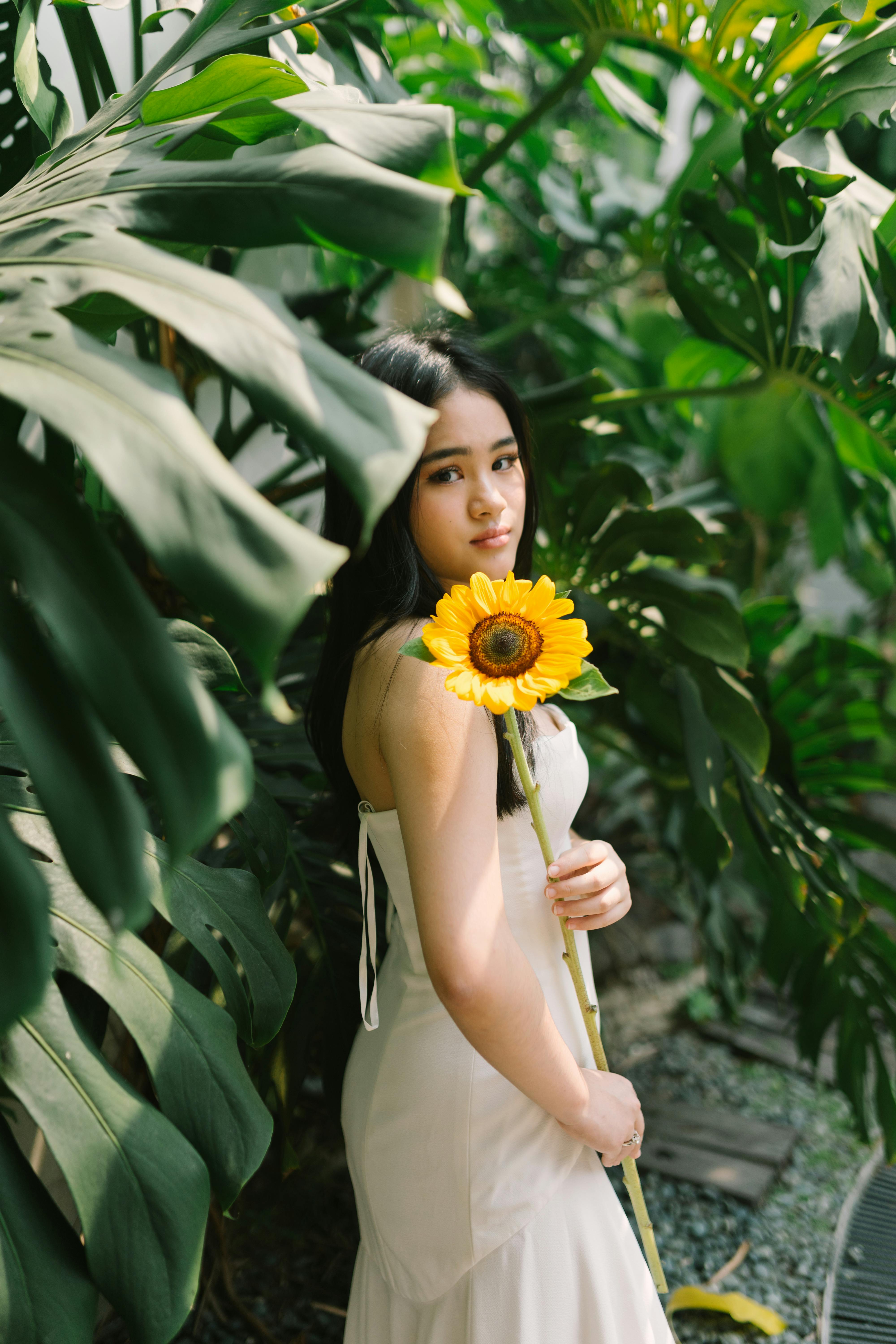 Young woman in white dress holds a sunflower amidst lush monstera leaves.