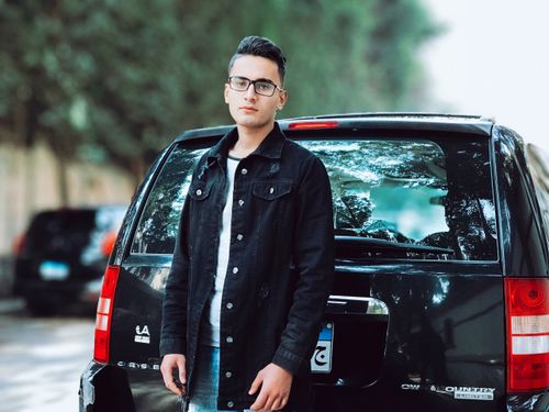 Free Young man in denim jacket posing confidently with a black SUV in an outdoor setting. Ideal for lifestyle and fashion concepts. Stock Photo