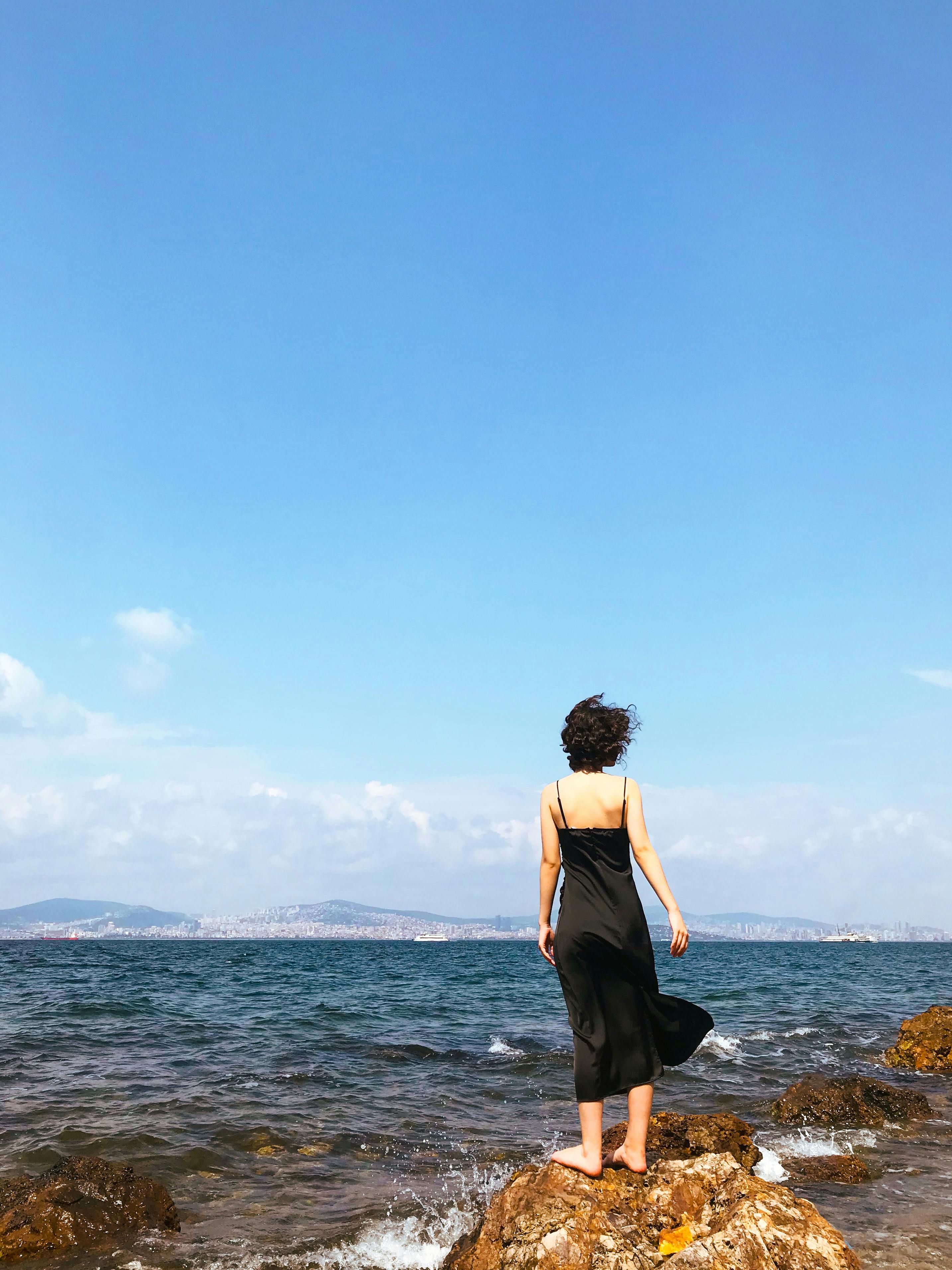 A woman in a black dress stands on a rocky shore, gazing at the sea in Burgazadası, Istanbul.