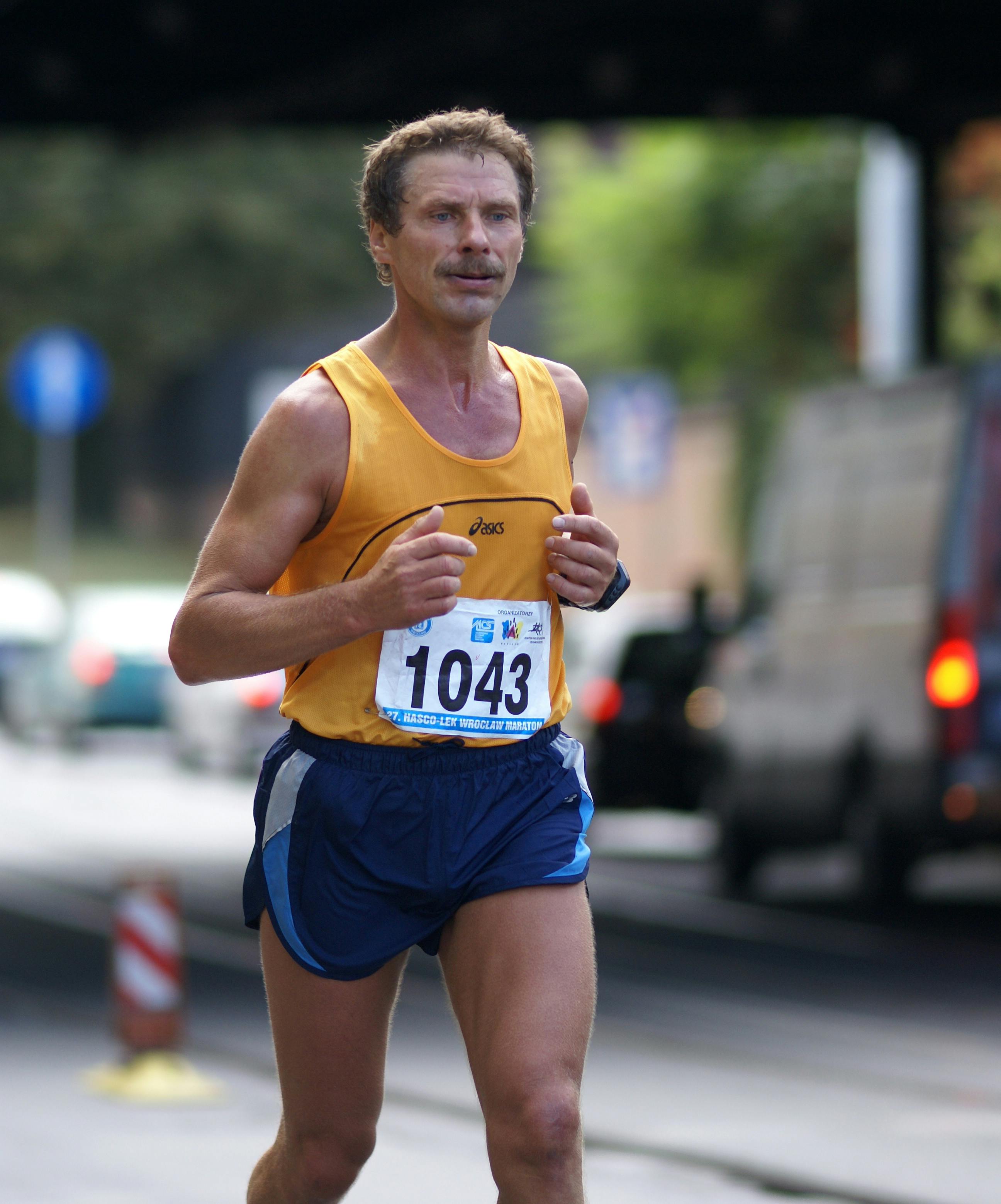 Middle-aged man running in a city marathon wearing race number 1043 on a bright day.