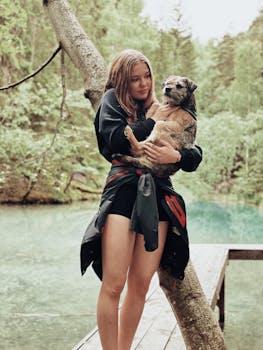 A young woman stands on a dock holding a dog, surrounded by a lush forest and clear lake.