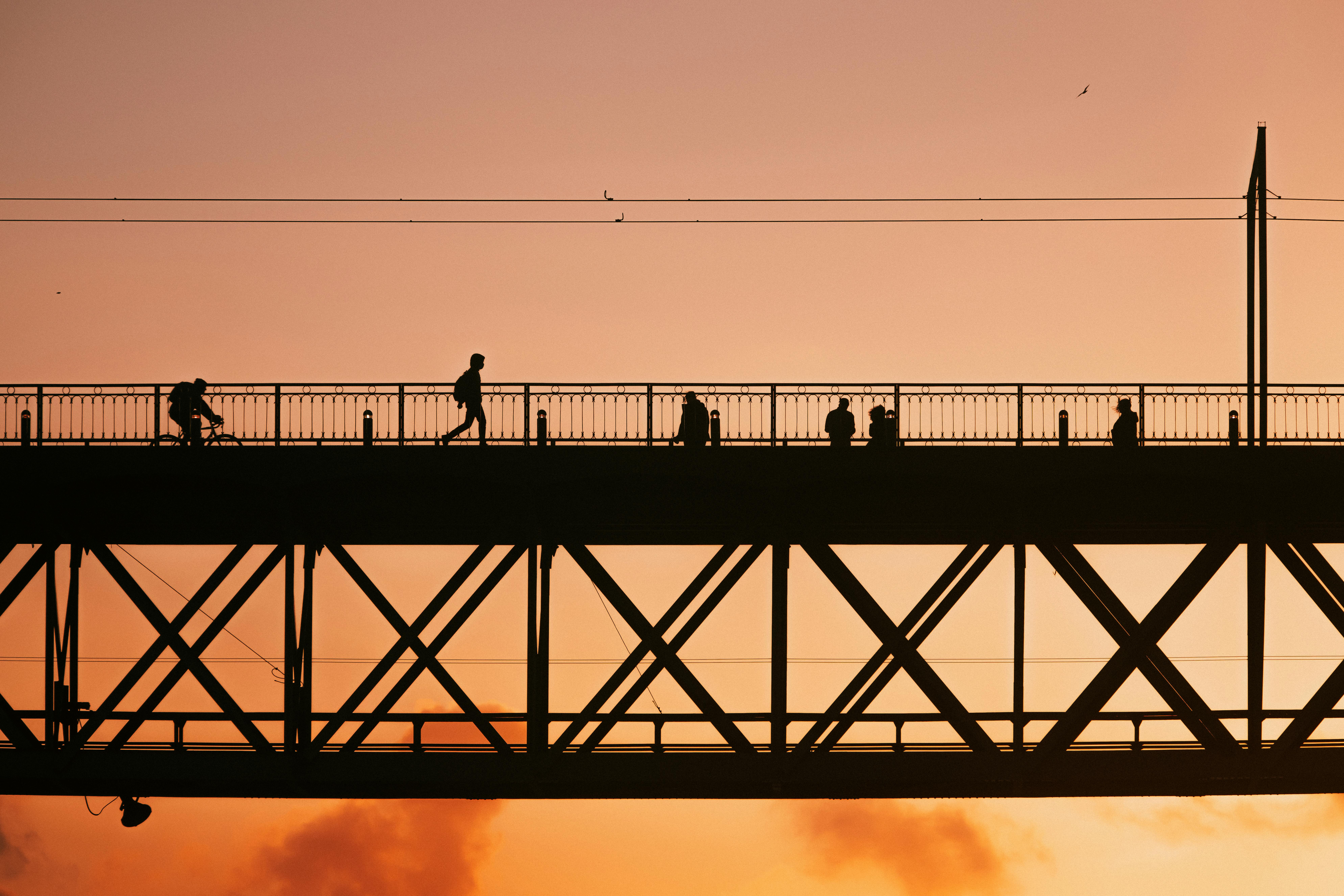 Silhouettes of people crossing a bridge against a warm sunset sky, showcasing urban life and architecture.
