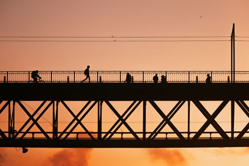 Free Silhouettes of people crossing a bridge against a warm sunset sky, showcasing urban life and architecture. Stock Photo